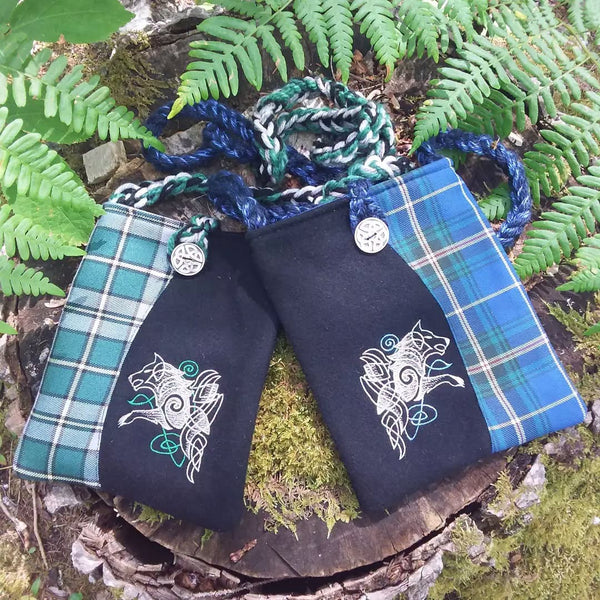Two small fabric pouches or bags with braided blue and green handles, placed on a mossy log surrounded by ferns. Each pouch has a tartan flap in teal and blue tones, a dark navy center panel, and white Celtic wolf embroidery.