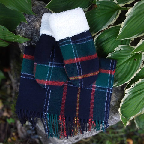 A pair of RCMP tartan mittens with a white faux-fur cuff, displayed on a rock surrounded by green hosta leaves. 